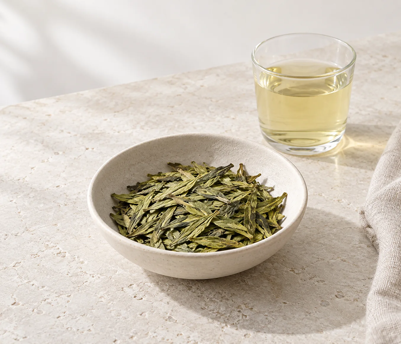 A shallow ceramic bowl of Longjing tea and a glass of pale amber tea on a light stone table